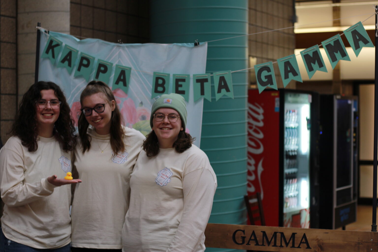 Three Lawrence Tech students stand in front of a teal banner reading 'Kappa Beta Gamma' during recruitment week; one sorority member holds a rubber duck.
