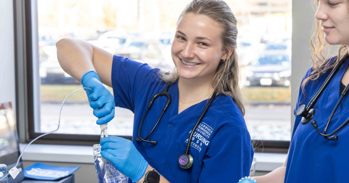 Smiling Lawrence Technological University nursing student in blue scrubs connects an IV tube to a fluid bag in the Nursing Sim Lab.